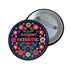 Round black pinback button with vibrant floral embroidery design surrounding bold pink and red text reading "Dissent is Patriotic."